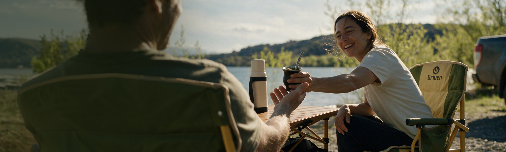 Dos personas se pasan el mate junto al agua, sentadas al aire libre